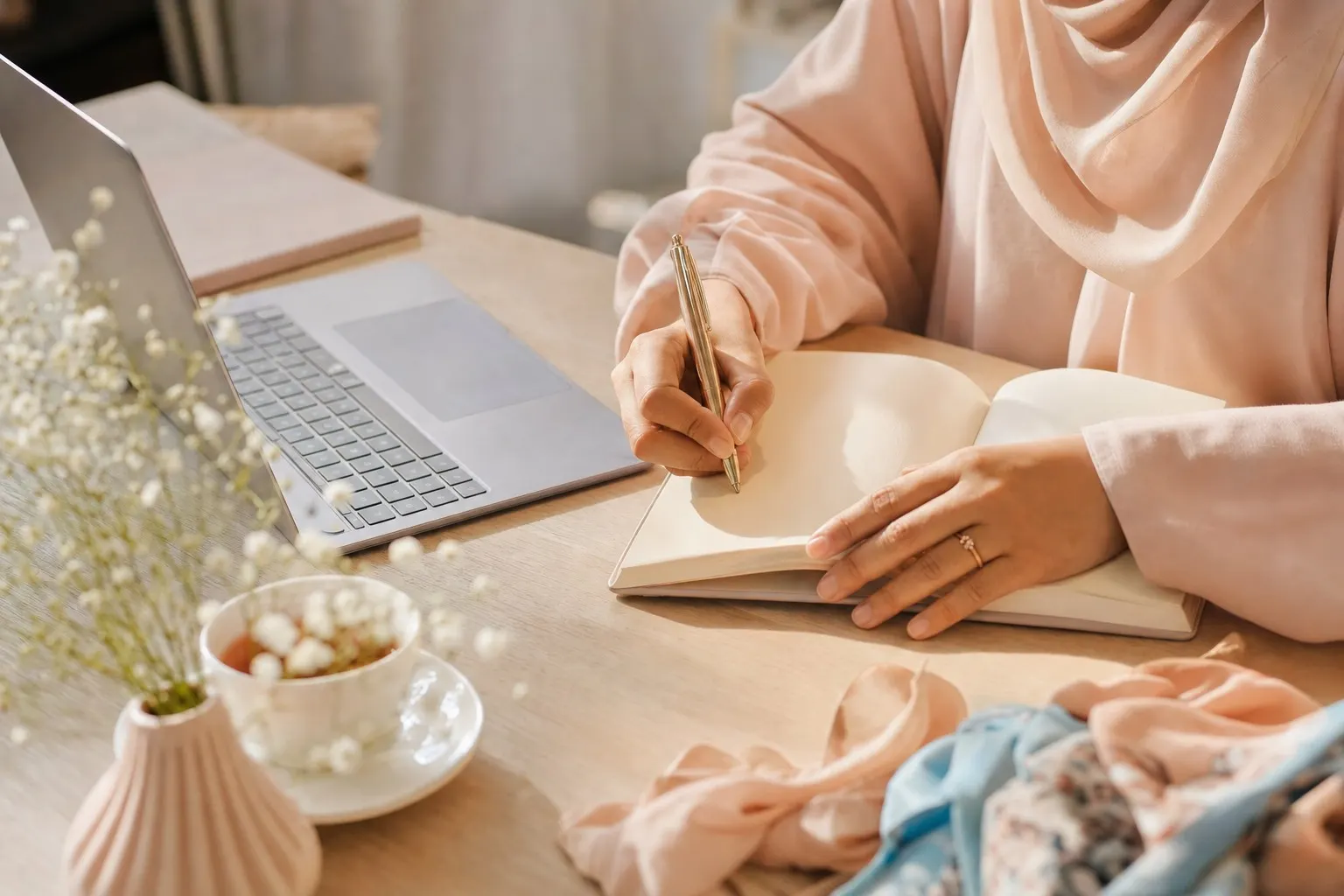 A woman’s hands writing in a cream journal beside an open laptop, with a cup of tea and small white flowers, in soft morning light